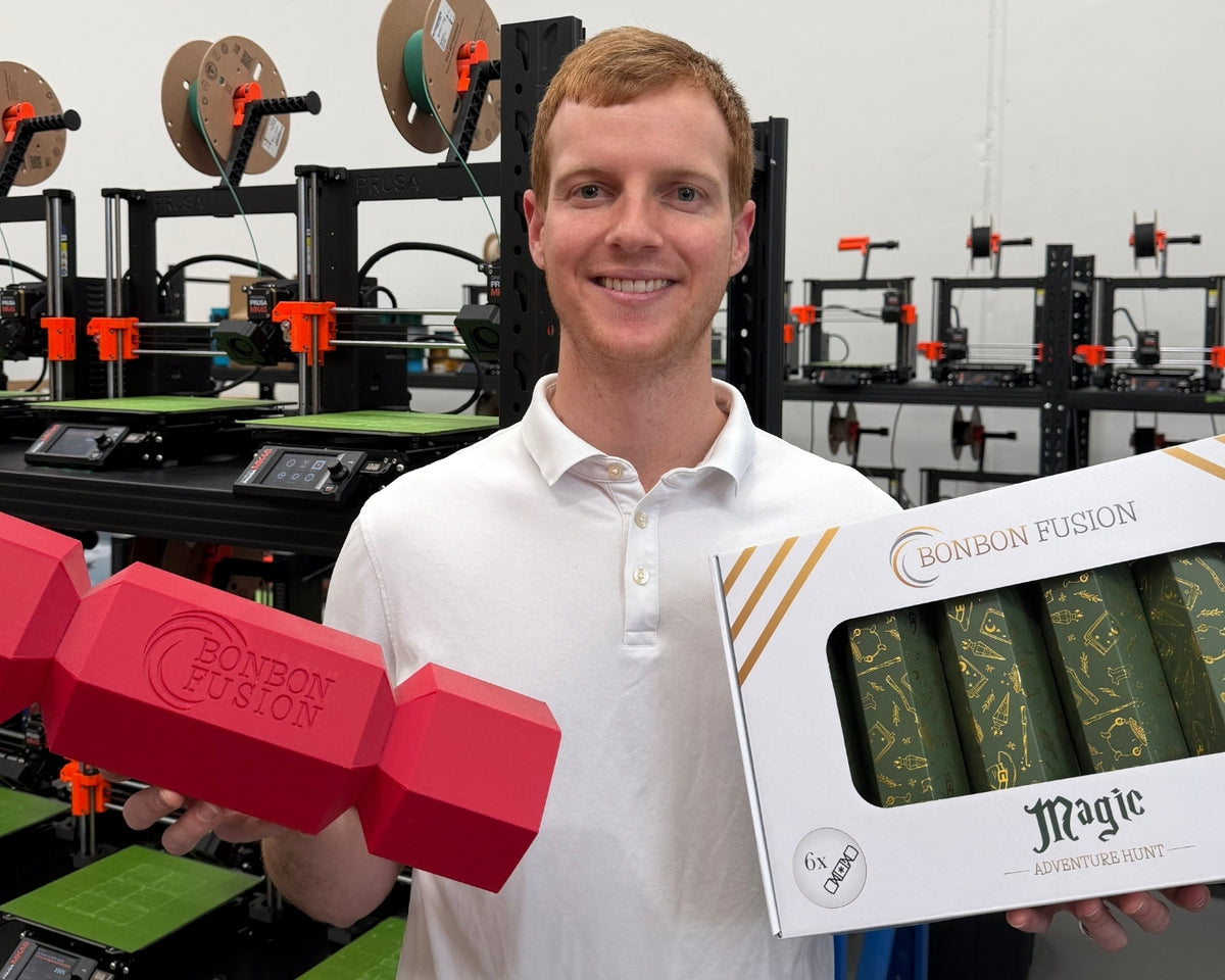 A smiling man in a white polo shirt holds a red Bonbon Fusion novelty item in one hand and a white Bonbon Fusion gift box containing green and gold wrapped items in the other, with 3D printers visible in the background.