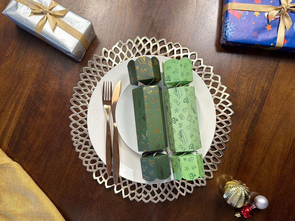 Two light and dark green Holiday Cruise Christmas bonbons on white plates with gold cutlery and a decorative gold placemat, surrounded by wrapped gifts and baubles on a wooden table.