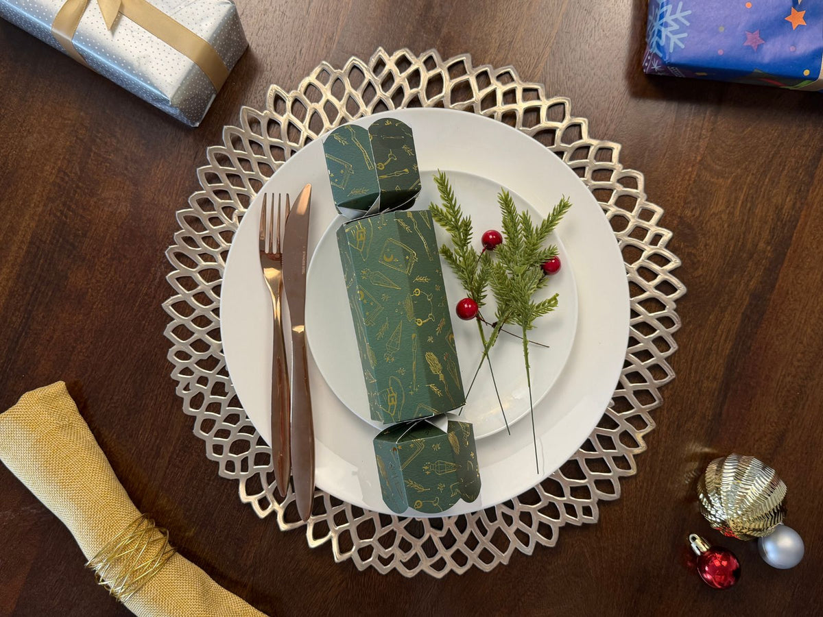 Bonbon Fusion Magic Christmas bonbon in green with gold foil magical illustrations on a white dinner plate, with rose gold cutlery, a gold napkin, a silver placemat, pine sprigs and red berries on a festive Christmas table