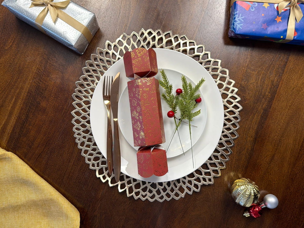 Bonbon Fusion Santa’s Workshop red and gold Christmas bonbon on a white plate with festive greenery, surrounded by gold cutlery, wrapped gifts and baubles on a wooden table.