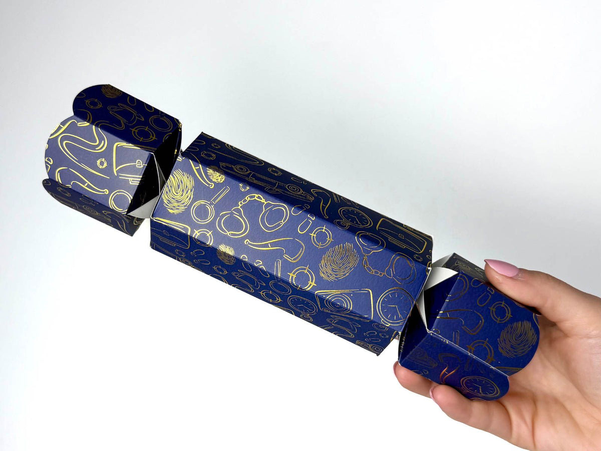 Hand holding a single Bonbon Fusion Spies Christmas bonbon in navy with gold foil spy illustrations, including fingerprints, handcuffs, magnifying glasses and pocket watches