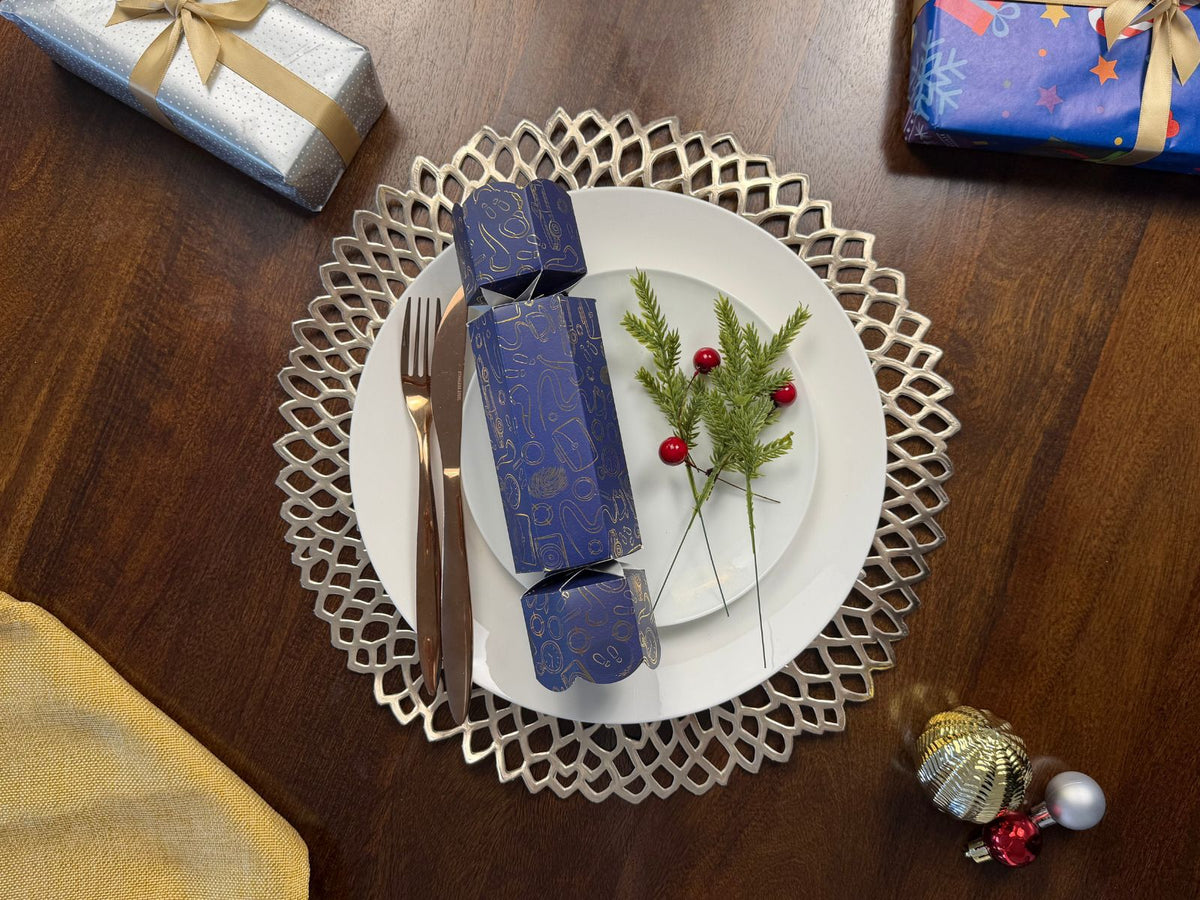 Bonbon Fusion Spies Christmas bonbon in navy and gold on a white dinner plate, set with rose gold cutlery, a silver placemat, pine sprigs and red berries on a festive Christmas table