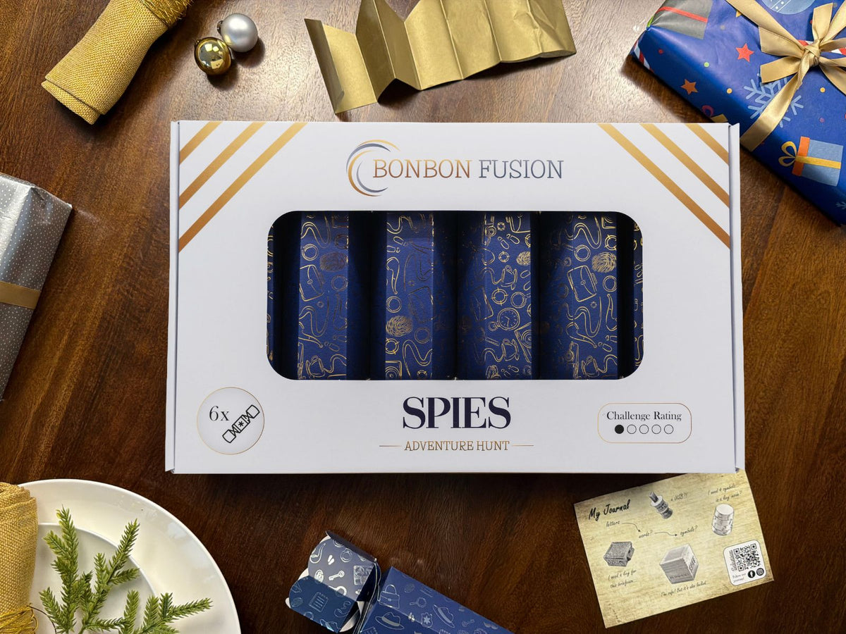 Bonbon Fusion Spies Adventure Hunt pack of six navy and gold Christmas bonbons on a festive dinner table, with a gold paper crown, a pulled bonbon and a "My Journal" puzzle clue card
