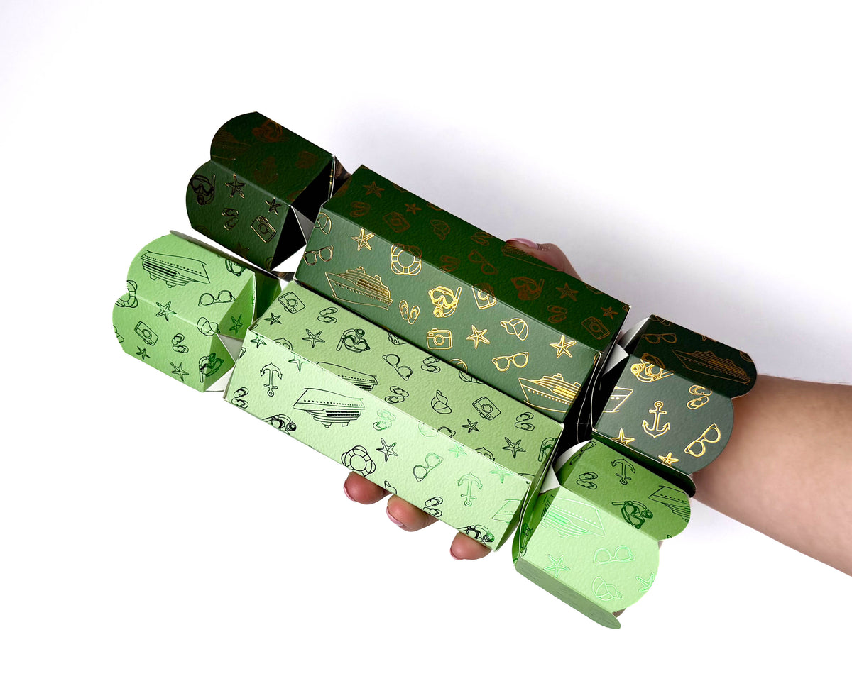 Hand holding two hexagonal Holiday Cruise Christmas bonbons in light and dark green, decorated with metallic nautical and holiday motifs, against a white background.
