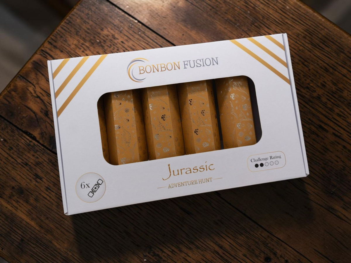 Bonbon Fusion Jurassic Adventure Hunt pack of six golden Christmas bonbons with dinosaur and fossil designs, in a white and gold windowed box on a wooden table.