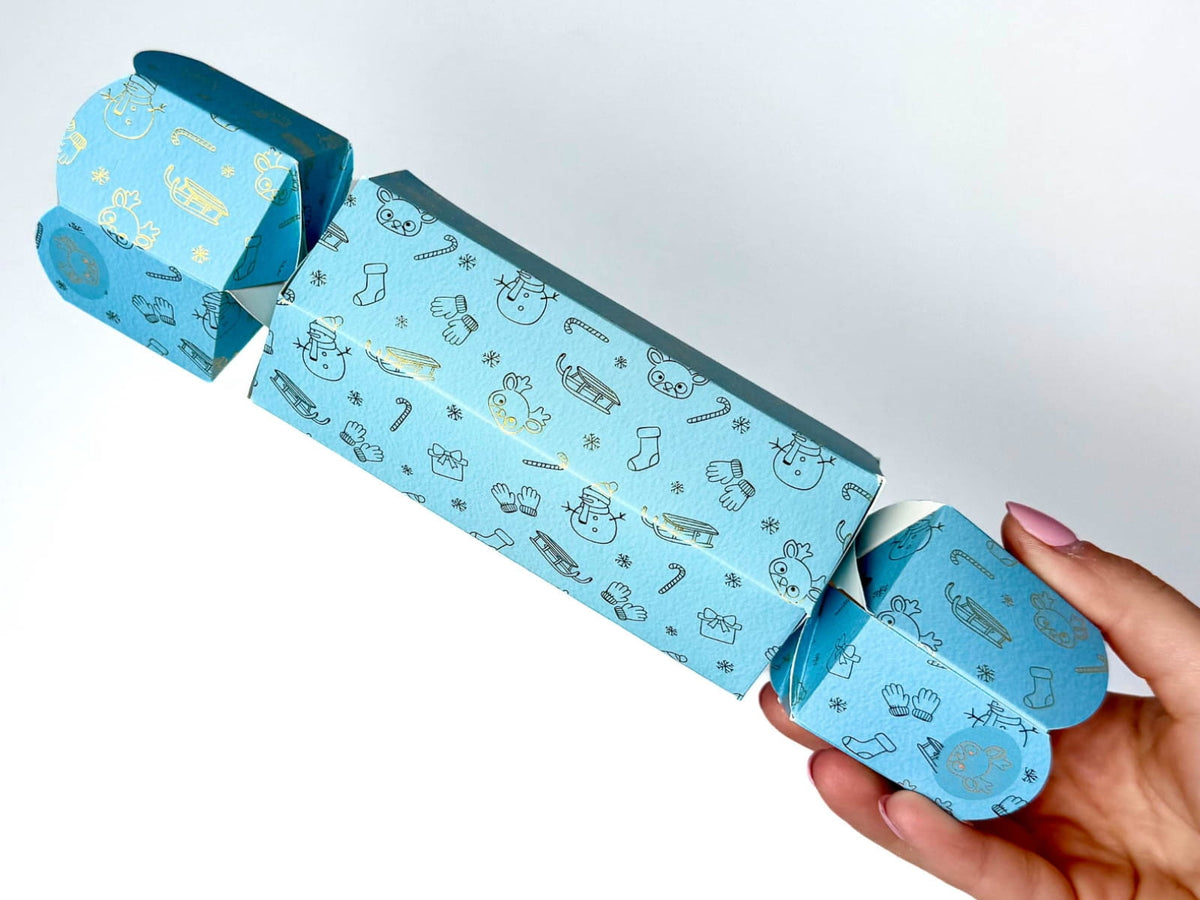 Hand holding a light blue hexagonal North Pole Christmas bonbon decorated with metallic festive designs, including snowmen, reindeer, sleighs and snowflakes, against a white background.