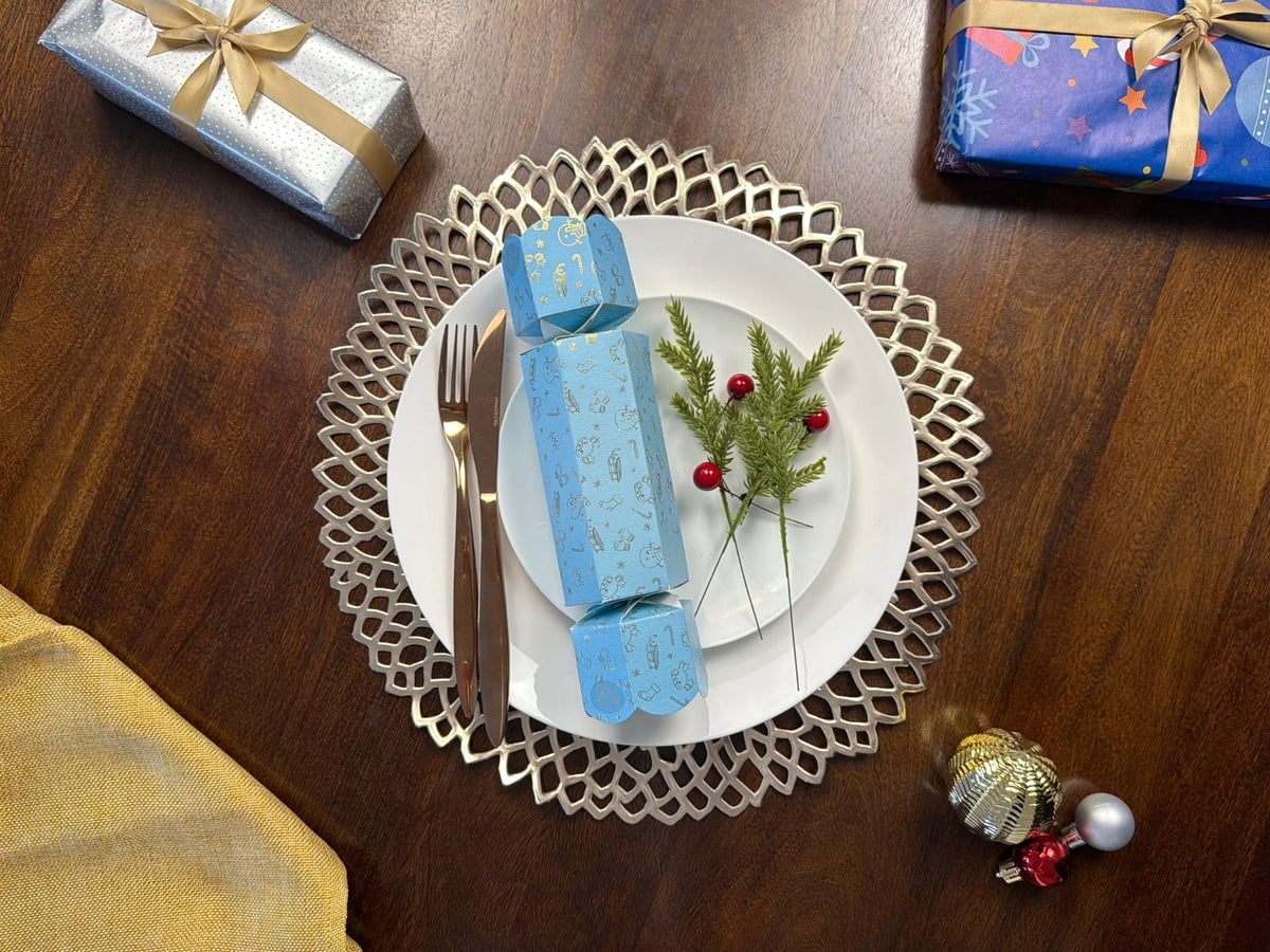 Light blue and gold North Pole Christmas bonbon on white plates beside greenery and red berries, with gold cutlery, a decorative gold placemat, wrapped gifts and baubles on a wooden table.