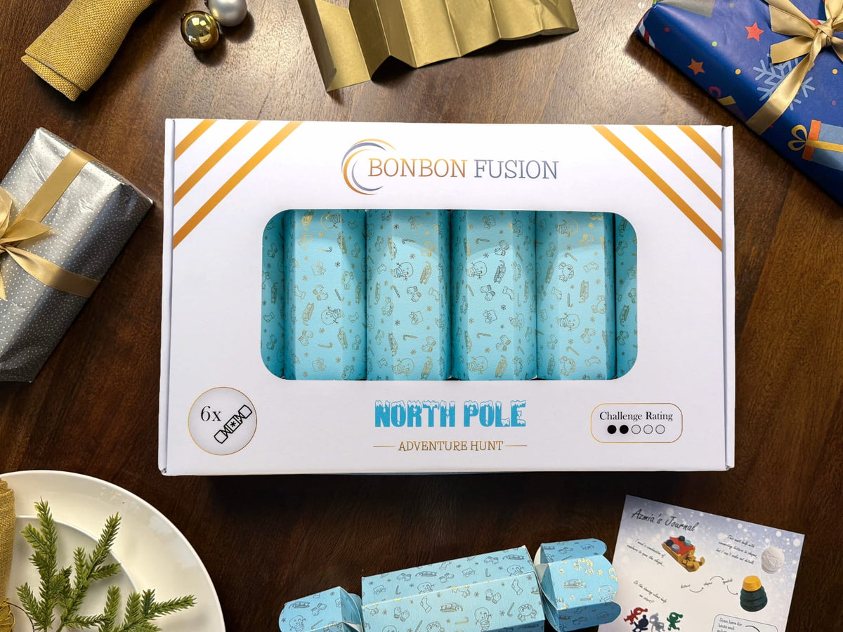 Bonbon Fusion North Pole Adventure Hunt pack of six light blue and gold Christmas bonbons on a wooden table, surrounded by wrapped gifts, a paper crown, Azmia’s journal and a festive place setting.