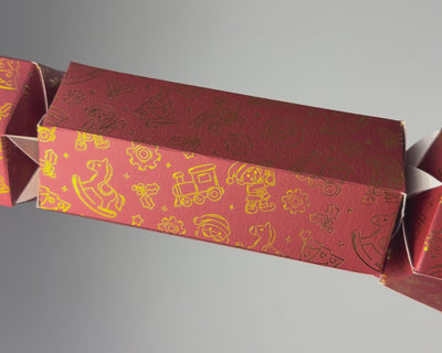 Hand holding a Bonbon Fusion Santa’s Workshop red Christmas bonbon decorated with gold illustrations of toys, elves, trains and Christmas trees against a white background.