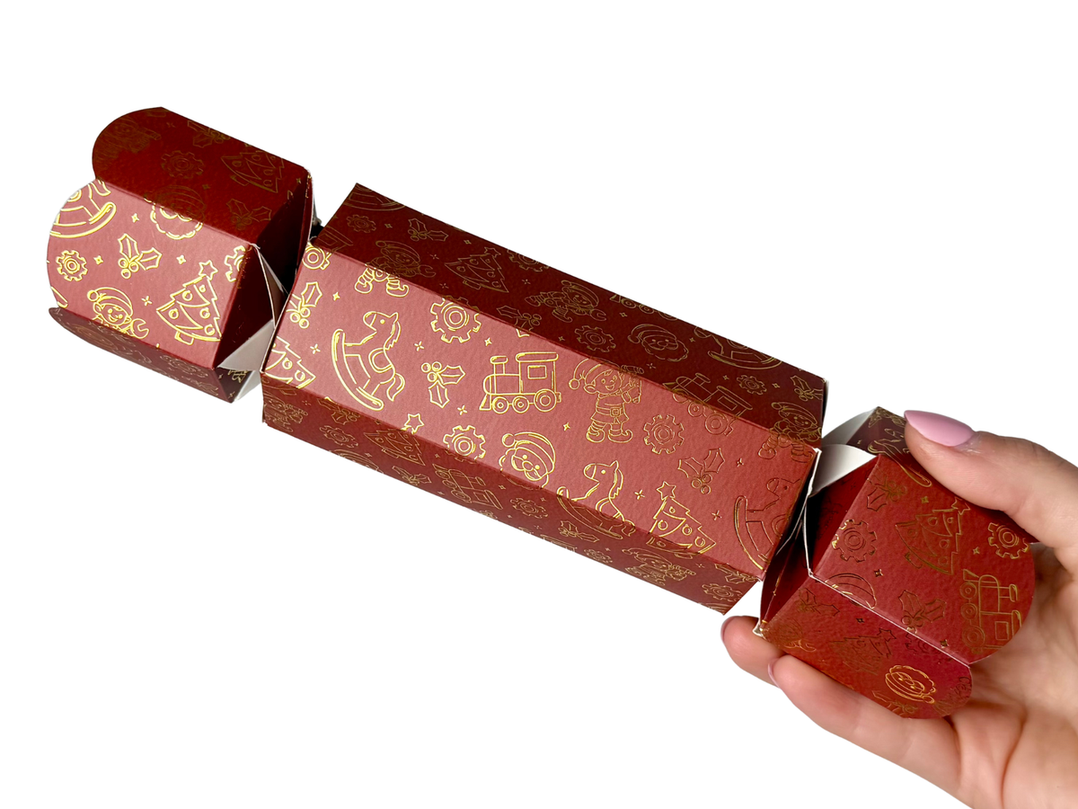 Hand holding a Bonbon Fusion Santa’s Workshop red Christmas bonbon decorated with gold illustrations of toys, elves, trains and Christmas trees against a white background.

